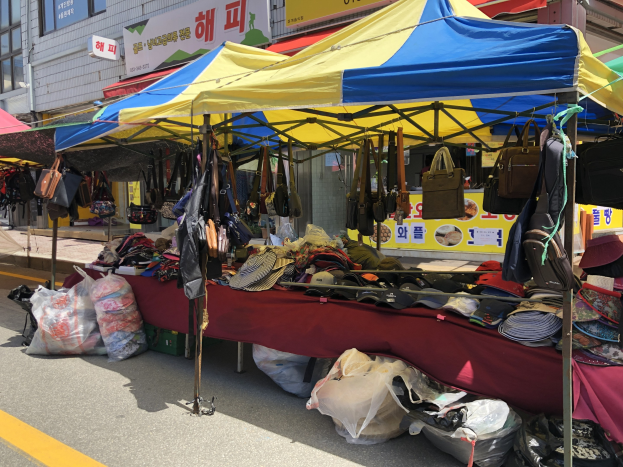 Ein Markt mit Zelten, die Taschen, Hüte und andere Gegenstände präsentieren, vor einem Hintergrund von Gebäuden mit Fenstern und Texttafeln.