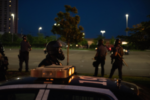 Eine Gruppe von Polizisten in Helmen mit Waffen vor einem markierten Polizeiauto in der Nacht zu sehen, mit Bäumen, Laternenpfählen, Gebäuden und einem sternenklaren Himmel im Hintergrund.