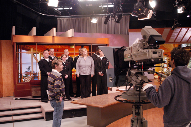 Gruppe von Menschen in einem Fernsehstudio mit einer Person, die eine Kamera hält, sowie an der Decke sichtbare Lichter.