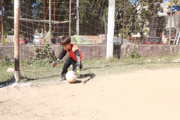 Ein junger Junge spielt Fußball auf einem Schmutzfeld mit einem bewölkten Himmel, umgeben von Gras, Pflanzen, Pfählen, einem Zaun, einer Wand, Bäumen, Fahrzeugen und Gebäuden.