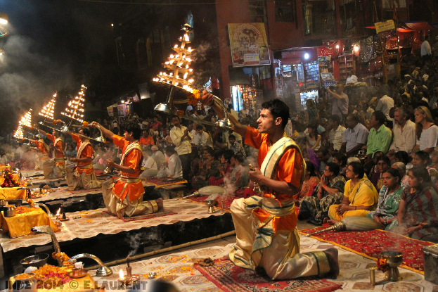 Eine Gruppe von Menschen in traditionellen Kostümen führt 'Aarti' in der Mitte durch, umgeben von traditionellen Gegenständen und Decken, mit Zuschauern, Gebäuden und verschiedenen Gegenständen im Hintergrund.