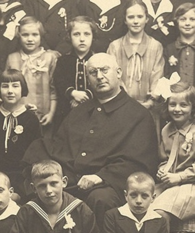 Ein altes Schwarz-Wei├č-Foto eines strengen Mannes, der in der Mitte sitzt und von einer Gruppe Kinder umgeben ist, die alle bewundernd zu ihm aufschauen.