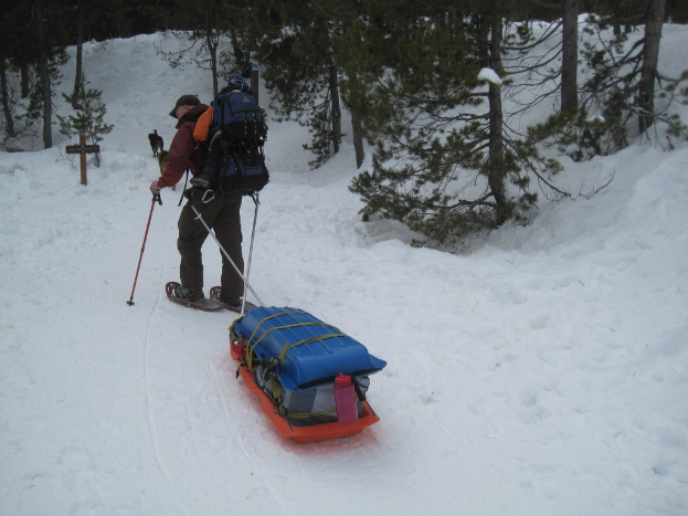 Ein Mann mit einer Tasche und Gepäck steht auf einem schneebedeckten Boden mit Bäumen im Hintergrund.