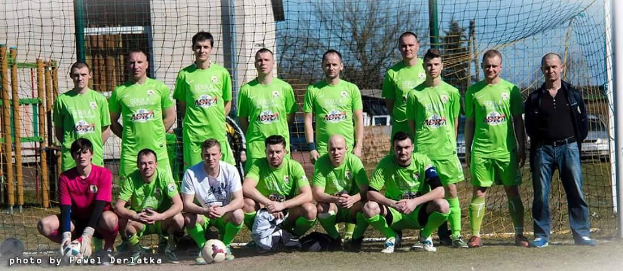 Mannergruppe in grünen T-Shirts auf einem Fußballfeld neben einem Torpfosten stehend, einige in Hockstellung, mit einem Ball davor und diversen Hintergrundelementen wie Bäume, Gebäude und Fahrzeuge.