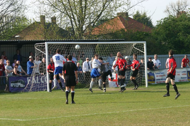 Spieler spielen Fussball auf einem Feld mit einem Tornetz, während Zuschauer dahinter stehen, mit Bäumen und Häusern im Hintergrund.