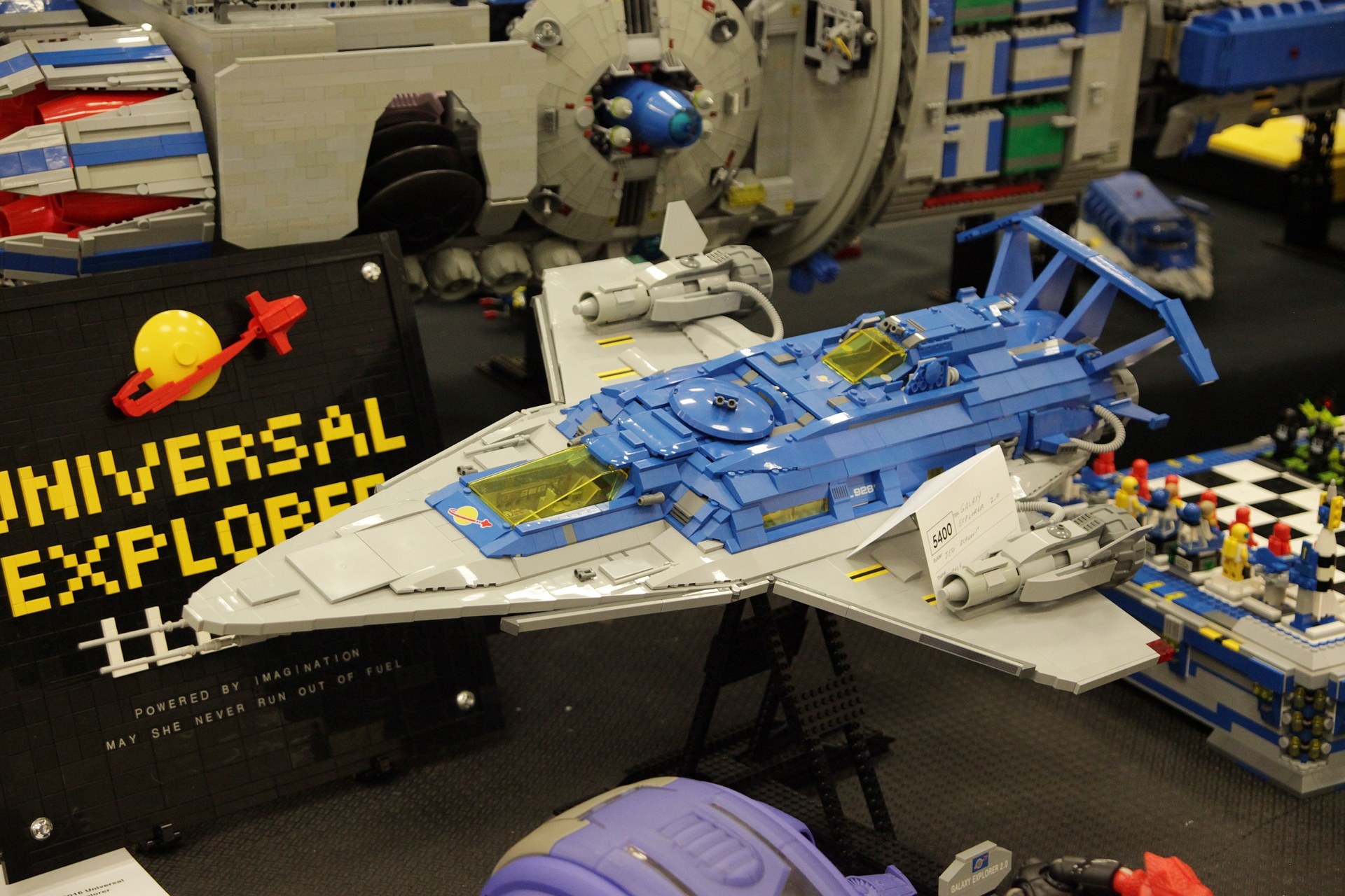 Ein Lego-Modell eines Raumschiffs auf einer Convention ausgestellt, umgeben von Spielzeugen und einer Tafel mit Text.