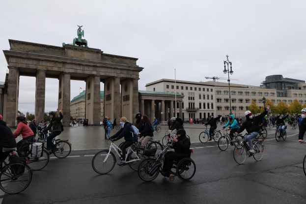 Eine Gruppe von Menschen auf Fahrr√§dern und Rollstühlen f√§hrt eine Stra√Ÿe entlang vor dem Brandenburger Tor in Berlin, Deutschland, mit dem Tor, das mit Säulen, einer Statue und Gebäuden mit Fenstern im Hintergrund geschmückt ist.