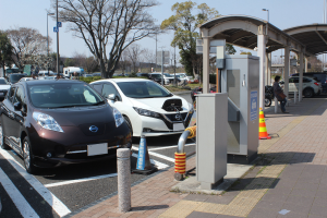 Elektroauto-Ladestation in Japan mit Autos, Verkehrskegeln, einer Person auf dem Gehweg, einem Schuppen, Polen, Lampen, Schildern, Bäumen, Pflanzen und unter einem Himmel.