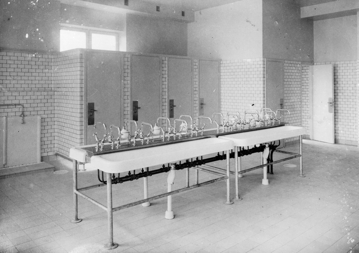 Schwarz-weiß-Foto einer öffentlichen Toilette mit einer Reihe Waschbecken, Wasserhähnen, Wänden, Türen, Fenstern und einem gefliesten Boden, mit verschiedenen Gegenständen, die herumliegen.