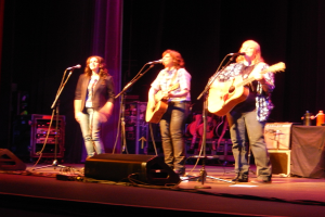 Drei Frauen treten auf der B├╝hne eines Musikkonzerts auf, jede singt in ein Mikrofon, waehrend sie eine Gitarre spielt, mit einem Tisch voller Wasserflaschen und Gitarren dahinter.