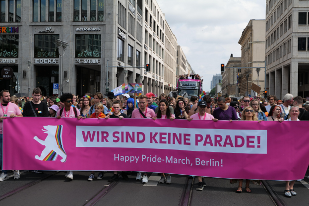 Gruppe von Menschen, die eine rosa 'Happy Pride March'-Fahne auf einer Straße in Berlin halten, mit Gebäuden, Laternenmasten und Verkehrszeichen an der Straße unter einem bewölkten Himmel.