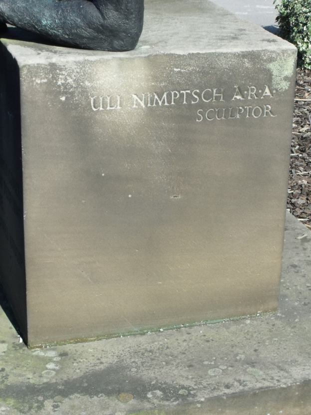Ein Stein mit Text in der Vordergrund, möglicherweise eine Statue oben, getrocknete Zweige, Erde und Pflanzen rechts, mit einer Straße im Hintergrund.