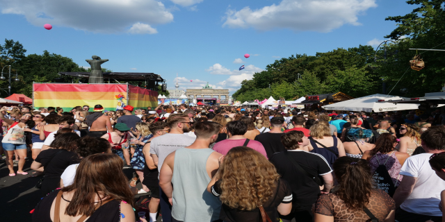 Eine große Menschenmenge, die eine Straße mit Zelten, Bäumen, Pfählen, Lichtern und einer Statue gesäumt, im Hintergrund Gebäude und ein wolkenverhangener Himmel mit Ballons, während des Christopher Street Day in Berlin.