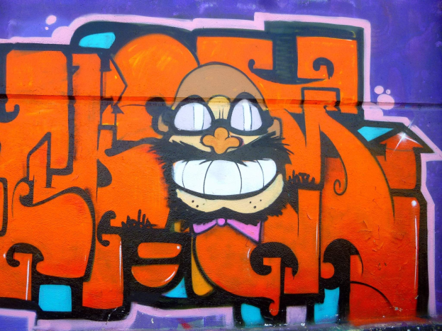 Graffiti-Gemälde eines Cartoon-Charakters mit großen Augen, einem breiten Grinsen und einem verschmitzten Gesichtsausdruck an der Seite eines Gebäudes, mit leuchtenden Farben und einer markanten Begleittext.