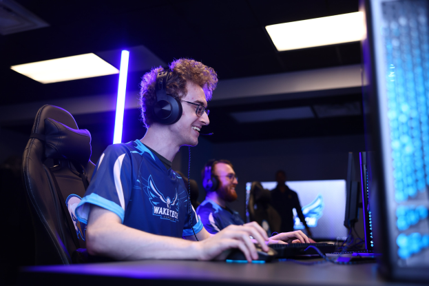 Ein Mann trägt einen Headset und Brille und sitzt an einem Schreibtisch mit einem Computer-Monitor, einer Tastatur und einer Maus, während er ein Spiel in einem Raum mit einer anderen Person und Deckenleuchten spielt.