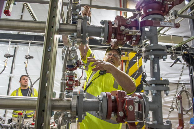 Zwei Männer in gelben Sicherheitswesten und Jacken arbeiten an einer Maschine in einer Fabrik mit Metallstangen, Rohren und einem weißen Tuch im Hintergrund; einer trägt eine Brille und eine Mütze.