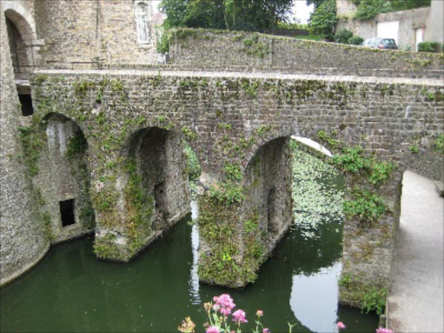 Eine Brücke spannt sich über das Bild, mit einer Straße, einem Baum und Wasser darunter, einem Auto und einer Wand in der oberen rechten Ecke und einem Gebäude auf der linken Seite.