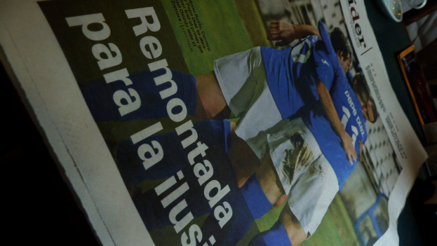 Eine Zeitung auf einem Tisch zeigt drei Männer in blauen Hemden, möglicherweise beim Fußballspielen, mit Text am unteren Bildrand.