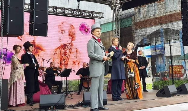 Eine Gruppe von Menschen, die auf einer Bühne stehen, mit einem Mann in der Mitte, der ein Mikrofon und ein Buch hölt, einer Frau, die links von ihm ein Musikinstrument spielt, und einem Mann, der rechts von ihm ein Blatt Papier hölt, während im Hintergrund jemand eine Geige spielt.