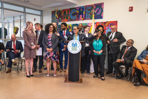 Gruppe von Menschen vor einem Podium mit einem Mikrofon und einer Tafel mit der Aufschrift "Sancho-Stadtrat tritt dem Stadtrat bei", mit sitzenden und stehenden Teilnehmern, Fahnen, einem Feuerlöscher und Deckenleuchten im Hintergrund.