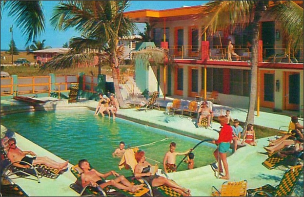 Ein Schwimmbad mit Menschen auf Stühlen im Vordergrund, Gebäuden, Bäumen und Autos im Hintergrund.