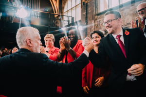 Ein Mann im Anzug und eine Frau in einem roten Kleid geben sich die Hand, während eine Gruppe applaudiert, mit Fenstern, Geländer und Lichtern im Hintergrund.