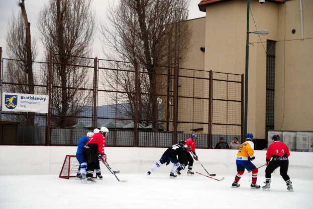 Menschen spielen Eishockey auf einem Eis mit Gebäuden, Bäumen, einer Straßenlaterne, einem Namensschild und Zäunen im Hintergrund bei klarem Himmel.