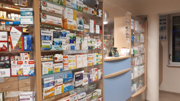 Eine Apotheken-Inneneinrichtung mit Regalen, die mit verschiedenen Medikamenten gefüllt sind, einer zentralen Säule und einem Glasfenster auf der rechten Seite, mit der Decke oben.
