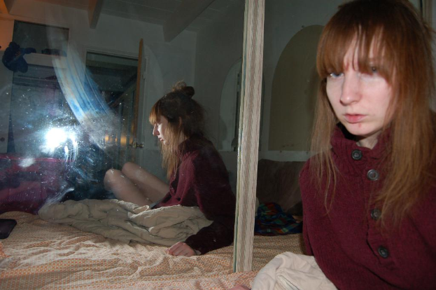 Eine Frau in einer roten Jacke steht neben einem Spiegel, der ihr Bild reflektiert, mit einem Bett und einer Bettwäsche im Hintergrund.