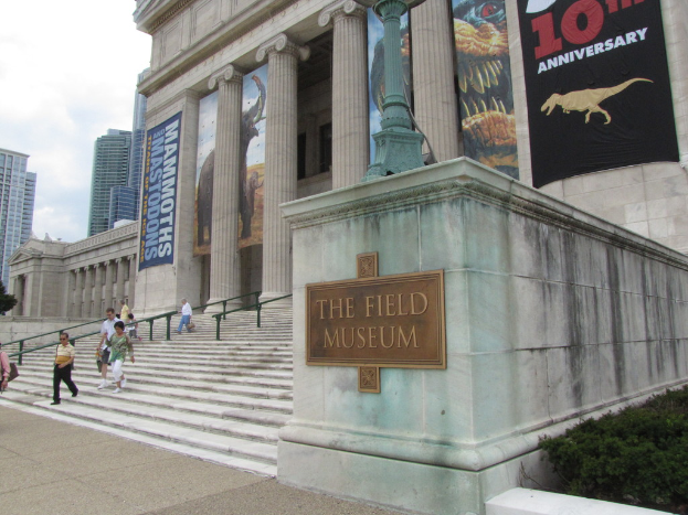 Menschen auf Treppenstufen neben einem Geländer sitzend, mit einem Denkmal auf der rechten Seite und Gebäuden im Hintergrund; ein Banner, Plakate und eine Säule sind oben zu sehen, ein Museumsschild in der Mitte und Planeten in der unteren rechten Ecke, mit Himmel und Wolken in der oberen linken Ecke.