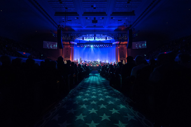 Ein großer Zuschauerraum mit Stühlen, eine Bühne mit Musikern und eine Decke mit Lichtern und Fernsehern, alles in einer dunklen Atmosphäre, die an einen Konzertsaal während der Nationalkonvention 2019 erinnert.