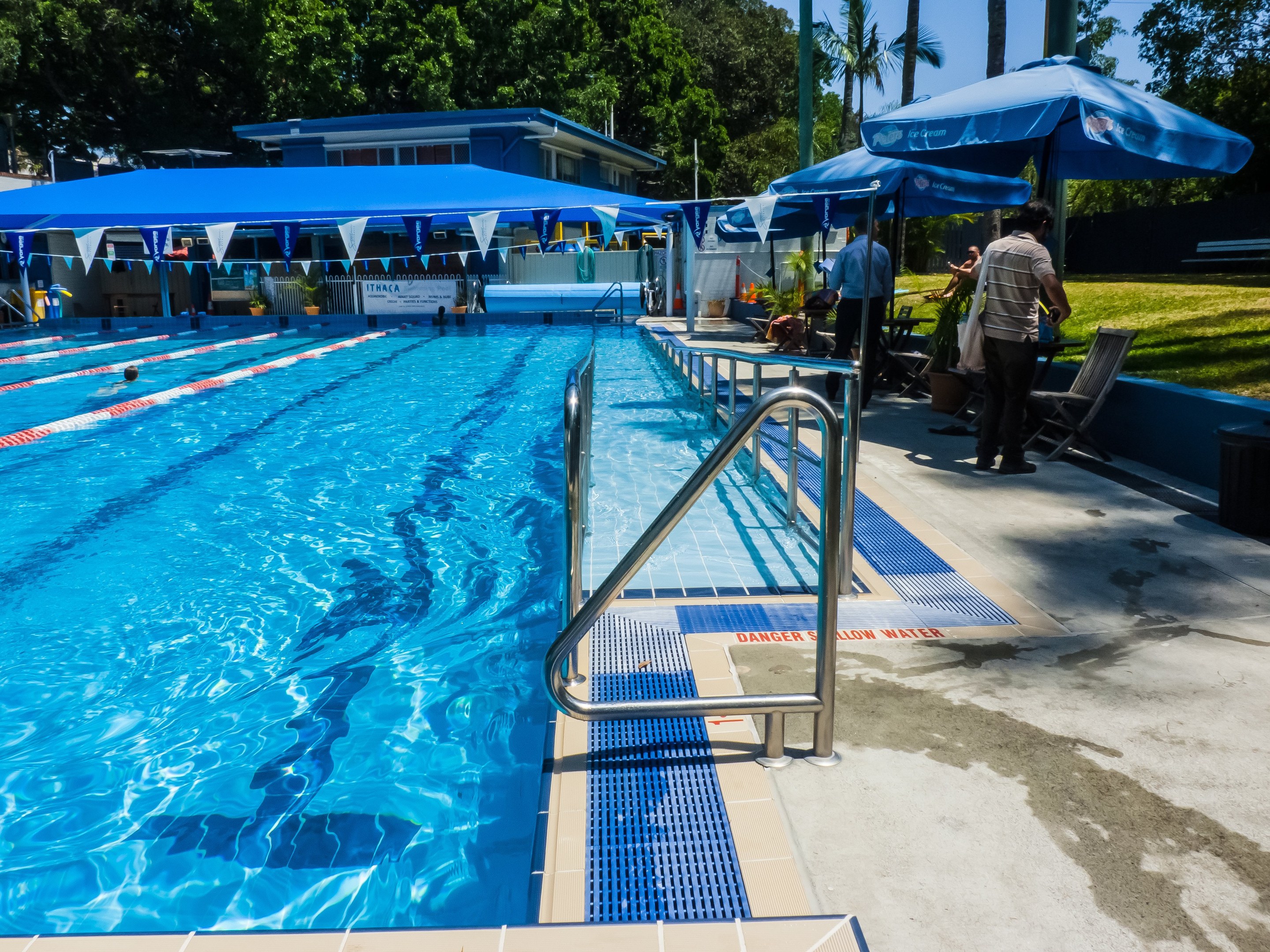 Großer Freibad mit schwimmenden Menschen, umgeben von Geländern, Sonnenschirmen, Stühlen, Tischen, Pflanzen, Gras, Bäumen und einem Gebäude unter einem klaren blauen Himmel.
