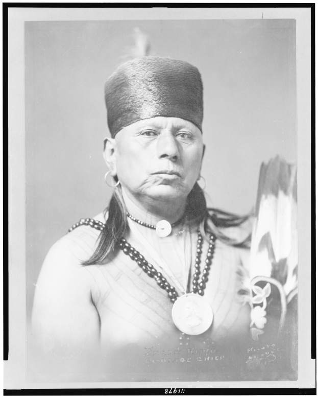 Ein altes Schwarz-Weiß-Foto eines Native American Man mit langen dunklen Haaren, traditioneller Kleidung und einer Mütze mit Verzierungen, der direkt in die Kamera schaut.
