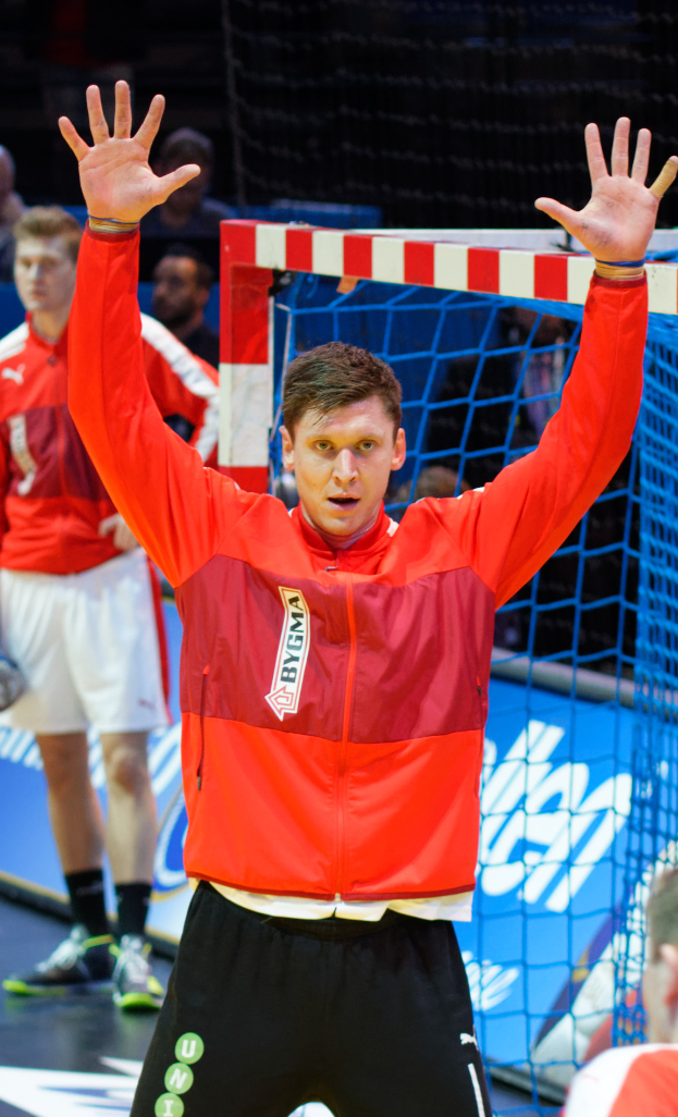 Ein Mann in einer roten Jacke und schwarzen Shorts steht auf einem Futsal-Platz, trägt Schuhe und hat ein Netz und einige andere Personen im Hintergrund.