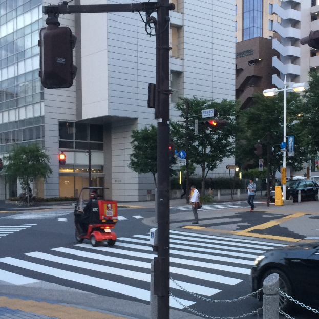 Ein Roller befindet sich auf der Straße mit einigen Personen dahinter, eine Ampel vorne und Gebäude mit Bäumen im Hintergrund.