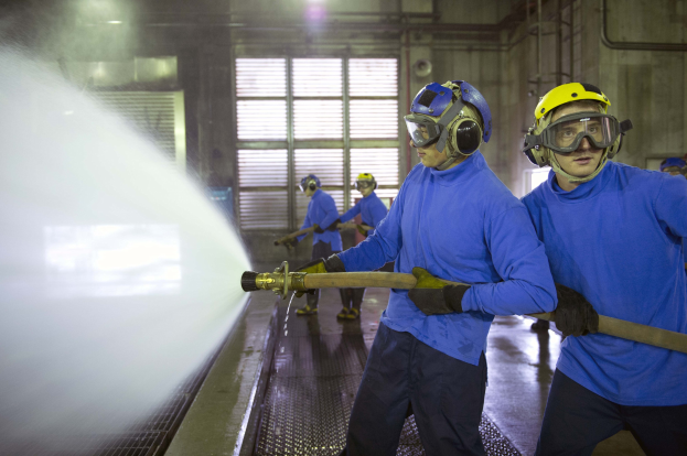 Gruppe von Männern in blauen Hemden und gelben Helmen, die an einer Maschine arbeiten, einer sprüht Wasser auf den Boden in einer Fabrikumgebung mit sichtbaren Wänden, Fenstern, Rohren und Lampen.