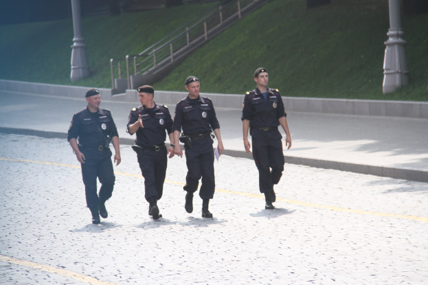 Drei Polizisten in Uniform und mit Mützen gehen eine Straße entlang mit Laternen, Geländern, Treppen und Gras im Hintergrund.
