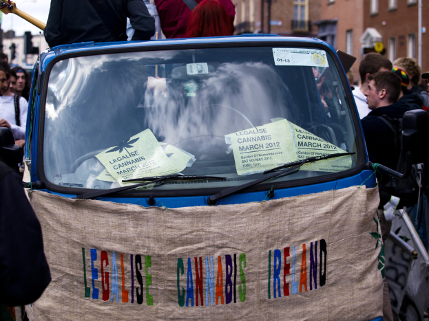 Eine Gruppe von Menschen steht um ein Auto herum, auf dem ein "Legalise Cannabis Ireland"-Schild steht, umgeben von Gebäuden mit Fenstern und Laternen unter einem klaren blauen Himmel, mit Papieren im Auto sichtbar.