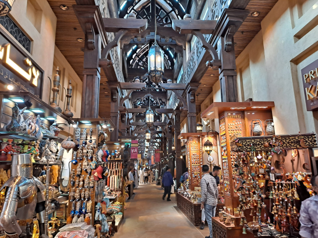 Ein geschäftiges Dubai-Basar mit Menschen, die zwischen Regalen voller Gegenstände hindurchgehen, Wänden mit Texttafeln und einer Decke mit warmem Licht.