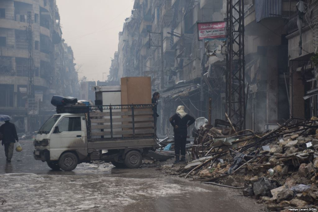 Ein Lastwagen fährt auf einer Straße neben einem Schutthaufen, während Menschen auf der Straße gehen, einer davon hält einen Schirm, und Gebäude, Pfosten und Schilder mit Text im Hintergrund unter einem klaren Himmel.