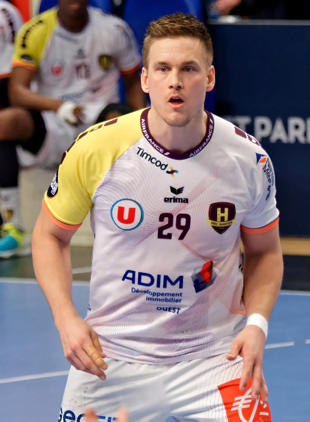 Ein Mann in einem weißen Hemd und gelben Shorts spielt Handball, hält ein Objekt und hat einen entschlossenen Gesichtsausdruck, vor einer Tafel mit der Aufschrift "Ligue 1 - Futsal - Olympique de Marseille" und einigen Menschen, die auf dem Boden sitzen.