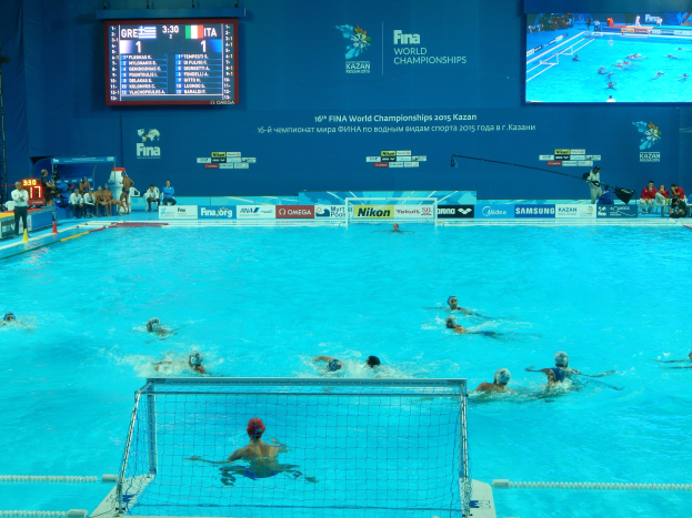 Wasserballspieler bei den 2016 FINA-Weltmeisterschaften in Kazan im Pool gegeneinander antretend, während auf der linken Seite Zuschauer sitzen und ein Anzeigeboard im Hintergrund die Veranstaltungsgestaltung anzeigt.