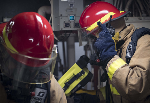 Zwei Feuerwehrleute in Schutzausrüstung bei der Arbeit an einem Feuerhydranten während einer übungsstunde mit Maschinen und Kabeln im Hintergrund.