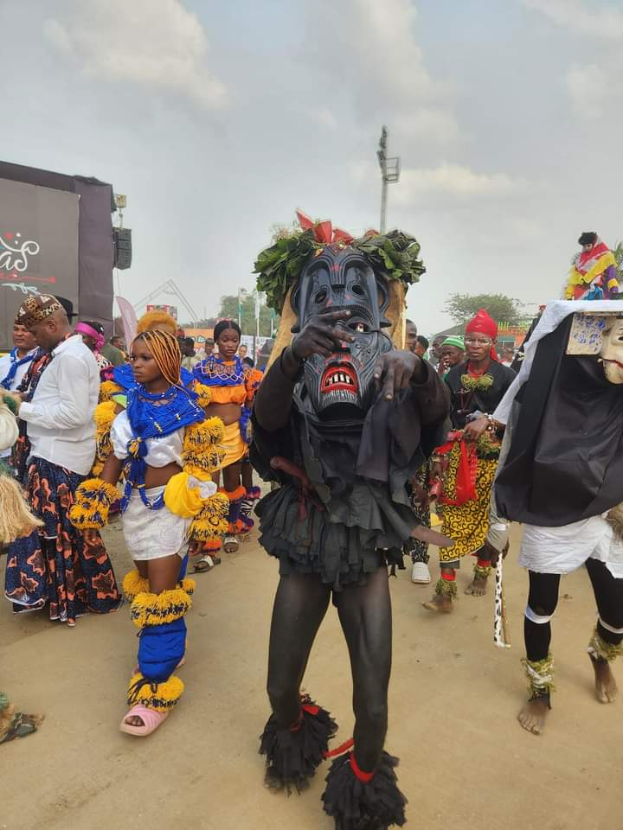 Eine Gruppe von Menschen in bunten Kostümen, die auf einer Straße während des Lagos Carnivals 2019 laufen, mit einer Person im Vordergrund, die eine Maske trägt und Bäumen, Pfählen und einem klaren blauen Himmel im Hintergrund.