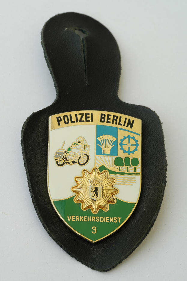 Ein schildförmiges Polizeibadged mit "Polizei Berlin" in schwarzer Fettschrift, das einen weißen Rand, einen blauen Hintergrund und einen zentralen weißen Stern aufweist und auf einer Oberfläche ruht.
