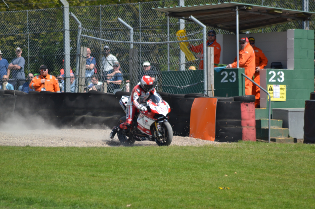 Ein Motorradfahrer mit Helm und Handschuhen fährt über ein grasbewachsenes Feld, mit Zuschauern in Mützen und Schutzbrillen, einem eingezäunten Bereich, Treppen mit Geländern und einem nummerierten Schuppen mit Texttafeln im Hintergrund.