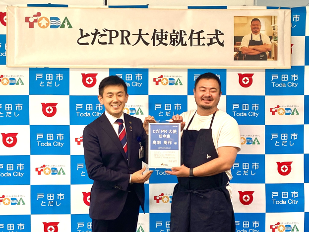 Zwei Männer mit einem Zertifikat, die lächeln, mit einem Banner "Toda City Wins PR Award" und einem Foto im Hintergrund.