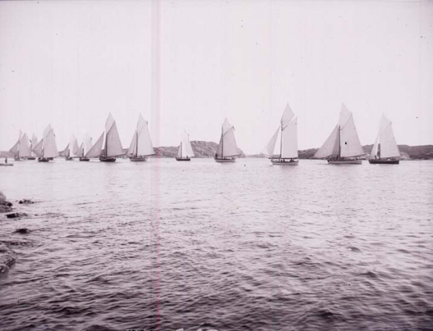 Schwarzes und weißes Foto mehrerer Segelboote auf dem Wasser mit Hügeln im Hintergrund und klarem Himmel, als Foto des New York Yacht Clubs bezeichnet.