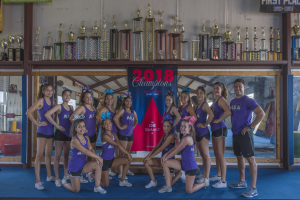 Gruppe von Cheerleadern in lila T-Shirts und schwarzen Shorts, die vor einer Vitrine mit Trophäen und einem Banner "2018 Summit Cheerleading Champions" posieren.