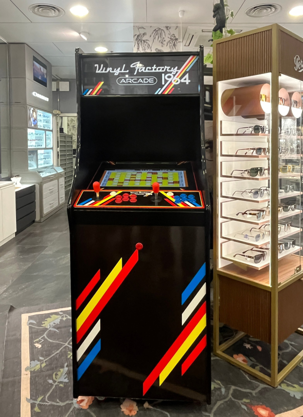 Ein Geschäft mit einem zentralen Vintage-Arcade-Automaten, einem Gestell mit Gläsern auf der rechten Seite und Hintergrundelementen wie Schränken, einem Fernseher und anderen Gegenständen, wobei Deckenleuchten für eine Retro-Atmosphäre sorgen.